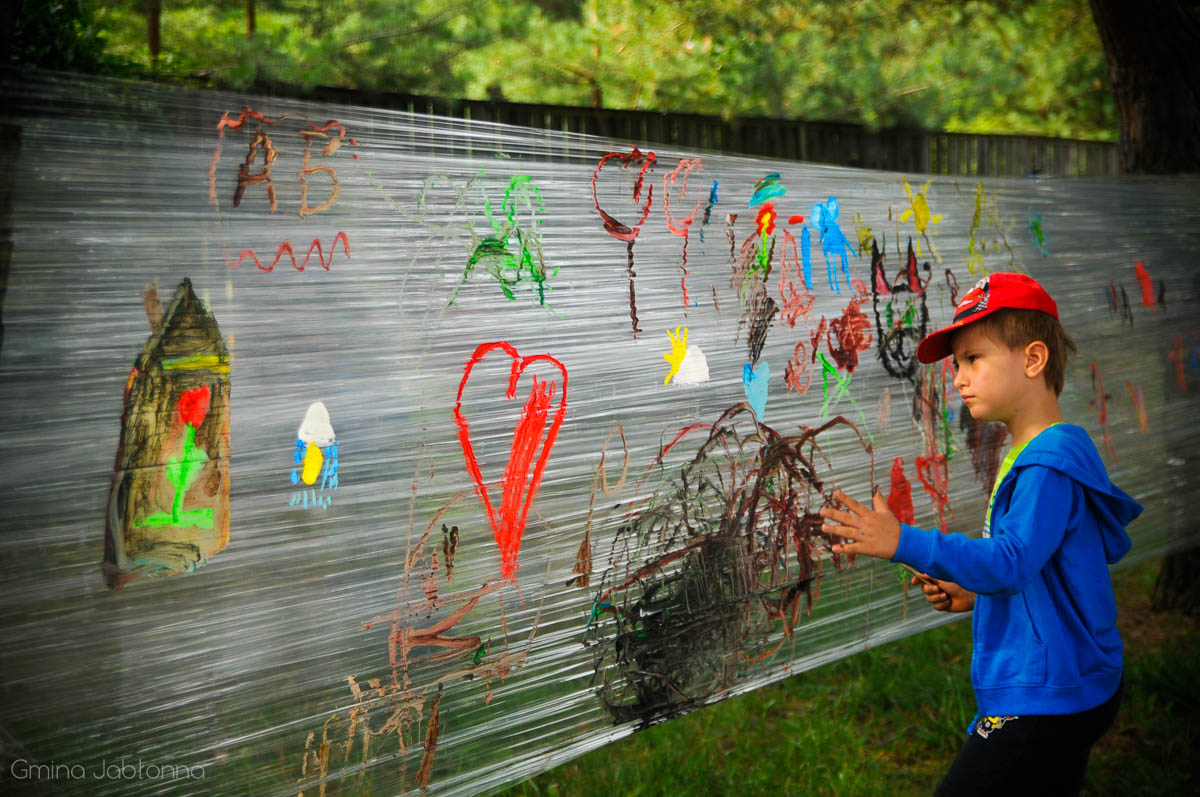 Chłopiec maluje kolorowymi farbami rysunki na przeźroczystej folii rozpiętej między drzewami. 